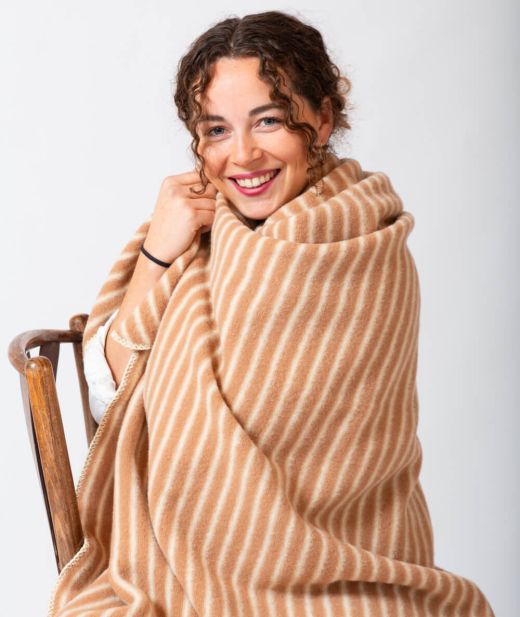 Femme assise enveloppée dans un plaid mérinos écru sable, lumière naturelle, ambiance chaleureuse.